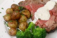 A piece of medium rare prime rib on a white plate with roasted baby potatoes and steamed broccoli.